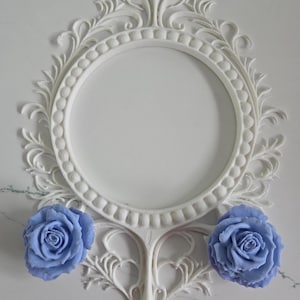 Puede incluir: Un marco de espejo ovalado blanco y ornamentado con un diseño floral detallado. Dos objetos en forma de rosa azul claro se colocan debajo del marco. El marco tiene un borde decorativo y texturizado.