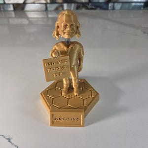 May include: A gold-colored bobblehead figurine of a person holding a sign that reads "DID YOU REBOOT IT?". The figurine is on a hexagonal base with the name "Bobbie Rob" printed on it. The head is spring-mounted.