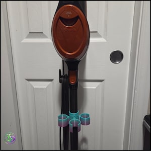 Peut inclure: Un aspirateur Shark noir et cuivre doté d'un manche vertical avec un cordon d'alimentation noir enroulé autour d'un crochet. Fixé au tube noir inférieur se trouve un support d'accessoires en plastique à trois branches, dégradé bleu sarcelle et violet.
