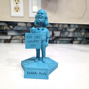 May include: A light blue bobblehead figure standing on a hexagonal base. The figure holds a sign that reads "DID YOU REBOOT IT?" and the base features a plaque with the text "Bobble-Tech". The figure has a spring neck and textured clothing.