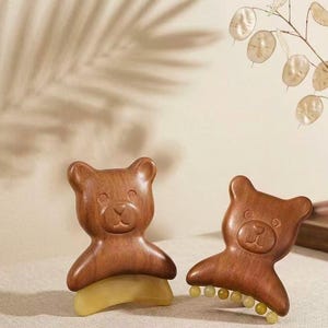 May include: Two bear-shaped wooden hair clips with smooth, dark brown finishes. One clip features a solid, light-coloured, translucent base, while the other clip has a row of small, round, light-coloured beads attached along the bottom edge.