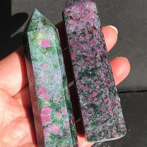 Peut inclure: Deux tours en cristal poli présentant un motif tacheté d'inclusions minérales magenta profond et vert forêt. Chaque pierre est taillée en prisme hexagonal avec un sommet pointu, offrant une surface lisse et réfléchissante aux variations naturelles.