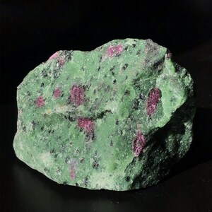 Könnte beinhalten: Ein rohes, unpoliertes Mineralexemplar mit einer leuchtend grünen Grundfarbe, eingebetteten tief magentafarbenen Kristallen und verstreuten schwarzen Mineraleinschlüssen auf der rauen, strukturierten Oberfläche des Steins.