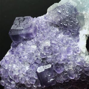 May include: A close-up of a fluorite mineral specimen. The crystal formation features a mix of purple and light blue hues, with a crystalline structure. The surface is covered in small, rounded formations and larger, cube-shaped crystals.