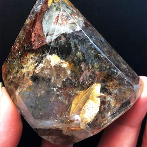 Pode incluir: Cristal transparente e facetado com inclusões douradas, castanhas e cinzentas. O cristal é segurado por uma mão, mostrando sua beleza natural e variações de cores. O cristal tem a forma de uma pirâmide truncada.
