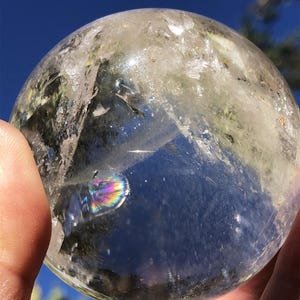 Peut inclure: Une boule de cristal de quartz sphérique et transparente présentant des fractures internes, des inclusions naturelles et une réfraction lumineuse aux couleurs de l'arc-en-ciel visible à l'intérieur de la pierre contre un ciel bleu.
