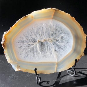 Può includere: Una fetta di agata traslucida di forma ovale con bande concentriche color crema, beige e bianco che circondano un centro cristallino bianco a forma di stella. La pietra è esposta su un supporto in metallo nero con due sostegni ricurvi.