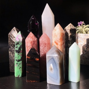 Peut inclure: Une collection de diverses tours en cristal poli aux couleurs et textures variées, incluant du vert et rose, brun foncé, blanc translucide, orange et gris à bandes, disposées en groupe sur un fond noir uni.