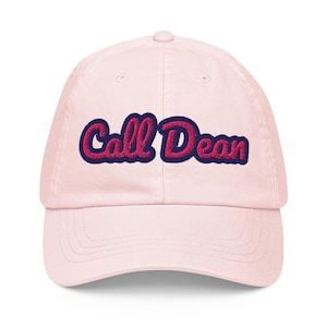 May include: A light pink baseball cap featuring the embroidered text Call Dean in a bold, cursive font. The lettering is colored in a vibrant magenta pink with a dark navy blue outline, centered on the front panel of the six-panel structured hat.