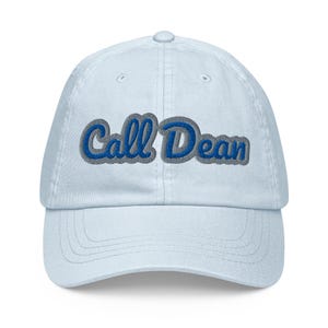 Puede incluir: Una gorra de béisbol azul claro con el texto bordado "Call Dean" en una fuente cursiva azul llamativa con un borde gris grueso. La gorra tiene una corona estructurada, una visera curva y un botón en la parte superior central de la estructura.