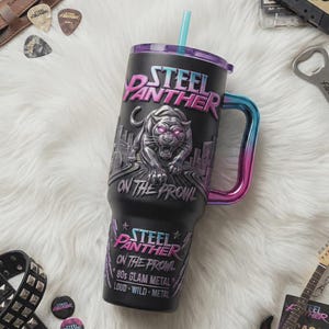 May include: Black tumbler with the text "STEEL PANTHER" and a panther graphic. The tumbler has a handle with a gradient of blue and pink. The text "ON THE PROWL" and "80s GLAM METAL" are also visible.