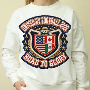 May include: White sweatshirt with a large embroidered patch. The patch features the text "UNITED BY FOOTBALL 2026" and "ROAD TO GLORY". The center design includes the flags of the USA, Canada, and Mexico, with the word "SOCCER" and the year 2026.