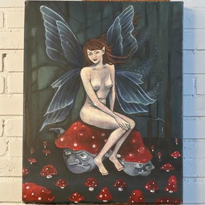 May include: An original painting depicting a fairy with blue wings and auburn hair, seated on a red mushroom with a face. The fairy is nude, set against a dark forest background with red mushrooms.