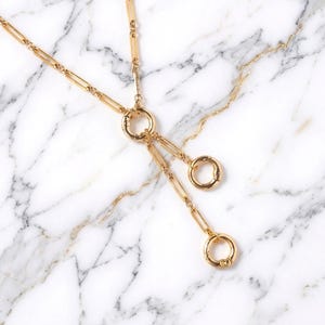 May include: A gold-toned necklace featuring a long chain with elongated rectangular links. The chain connects to a central circular spring ring clasp, from which two additional short chain segments hang, each ending in its own circular spring ring clasp.