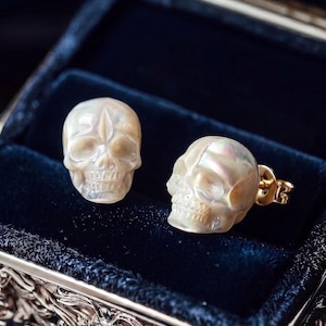 May include: A pair of stud earrings featuring carved mother-of-pearl skulls with iridescent surfaces. Each skull is detailed with defined eye sockets, a nasal cavity, and teeth, attached to gold-toned metal posts with secure backings.