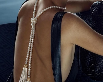May include: A long pearl necklace worn down the back, featuring a central knot detail and two dangling pearl tassels capped with gold-toned metal hardware. The necklace is paired with a black sleeveless garment and a small silver-toned earring.