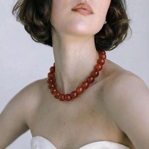 May include: A necklace composed of uniform, round, translucent reddish-orange beads. The beads are strung together in a single strand that rests around the neck, featuring a smooth, polished surface that reflects light across the circular shapes.