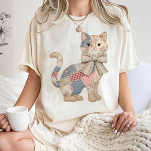 May include: Cream-colored t-shirt featuring a patchwork cat design. The cat has a bow tie and is made of various patterned fabrics, including floral, polka dot, and plaid. The shirt is a relaxed fit.