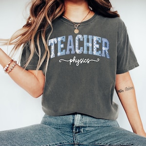 May include: Dark gray t-shirt with the word "TEACHER" in blue floral and gingham print letters. Below the word "TEACHER" is the word "physics" in a cursive font. The shirt is a casual style.