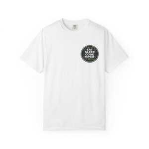 Può includere: T-shirt bianca con girocollo e maniche corte. Una grafica circolare sul petto recita "EAT SLEEP CODE REPEAT" in testo bianco e verde, con simboli di codice. La maglietta è realizzata in un materiale morbido.