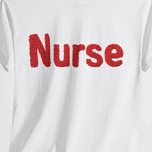 Può includere: T-shirt bianca con la scritta "Nurse" in lettere rosse e strutturate. La maglietta ha maniche corte e girocollo. Il design è centrato sulla parte anteriore.