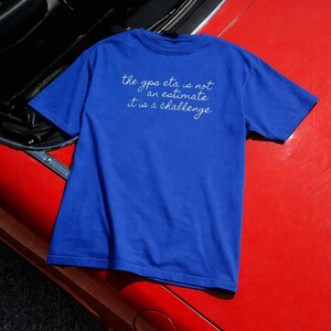 Peut inclure: Un t-shirt bleu roi avec le texte manuscrit "the gps eta is not an estimate it is a challenge". Le t-shirt est posé sur le capot rouge d'une voiture classique.