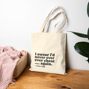 May include: Cream-colored tote bag with black text that reads "i swear i'd never ever ever cheat .... again." The bag has two handles and is made of a canvas-like material. The text is in a bold, sans-serif font. The bag is sitting on a wooden surface.