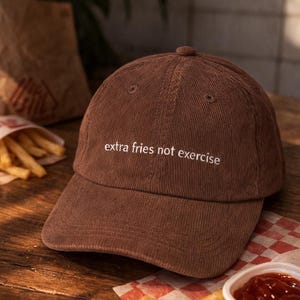 May include: Brown corduroy baseball cap with the text "extra fries not exercise" embroidered in white. The cap is on a wooden surface with fries and ketchup.