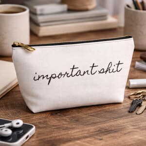 Op de afbeelding: Een crèmekleurige canvas make-up tas met de geborduurde zin "important shit" in zwart schrift. De tas heeft een gouden rits en ligt op een houten oppervlak met sleutels, een telefoon en een notitieboek.