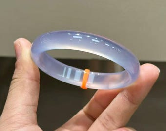 May include: A translucent, light blue jade bangle bracelet. The smooth, polished surface of the bracelet is accented by a thin, bright orange band. The bracelet is held in a hand, showcasing its elegant design and color.