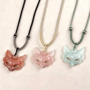 May include: Three necklaces featuring carved fox head pendants made of translucent stone in shades of reddish-brown, pale pink, and light green. Each pendant hangs from a matching adjustable cord with small sliding beads for length adjustment.