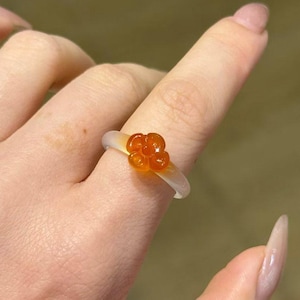 May include: A clear resin ring with a cluster of orange beads. The ring is worn on a finger, showcasing the translucent band and the vibrant orange beads. The ring's design is simple yet eye-catching.