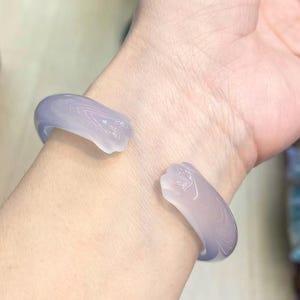 May include: A light purple open cuff bracelet. The bracelet is made of a translucent material with a marbled effect. The bracelet is worn on a wrist.