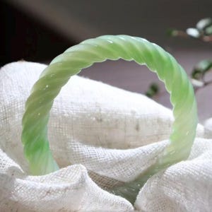 May include: A light green, twisted jade bangle bracelet. The bracelet has a spiral design and is resting on a white, textured fabric. The jade has a translucent quality, and the overall aesthetic is elegant and refined.