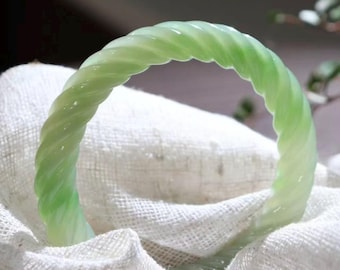 May include: A light green, twisted jade bangle bracelet. The bracelet has a spiral design and is resting on a white, textured fabric. The jade has a translucent quality, and the overall aesthetic is elegant and refined.