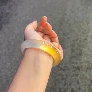 以下が含まれることがあります： 淡い白色から鮮やかな黄色へとグラデーションする、幅広の透明なバングルブレスレット。滑らかで磨かれた表面が光を反射し、ブレスレットのエレガントなデザインを際立たせています。