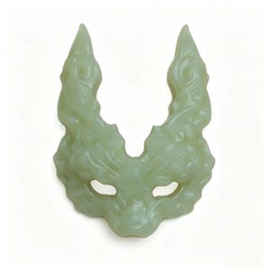 May include: A pale green mask with a stylized wolf or mythical creature design. The mask has two eye holes and two pointed protrusions at the top. The surface has a textured, carved appearance.