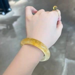 Könnte beinhalten: Ein durchscheinendes gelb-oranges Armreif-Armband mit glatter, polierter Oberfläche. Das Armband wird am Handgelenk getragen, und ein goldener Ring mit einem gelben Edelstein befindet sich an einem Finger. Der Schmuck befindet sich vor einem neutralen Hintergrund.