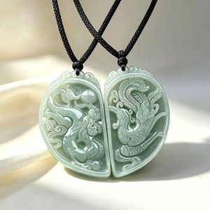 May include: Two pale green jade pendants shaped as halves of a heart, suspended on black cords. One pendant features a carved dragon, while the other displays a carved phoenix. The intricate relief designs are polished and light green in color.