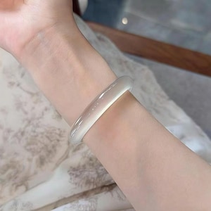 May include: A translucent, light-colored bangle bracelet. The smooth, rounded bracelet is worn on a wrist, showcasing its simple, elegant design. The bracelet appears to be made of a polished, possibly stone-like material, reflecting light with a subtle glow.