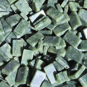 May include: A pile of green jade rectangular beads with rounded edges. The beads vary in shade from light to dark green and have a natural, slightly mottled appearance. Some beads have drilled holes for stringing.