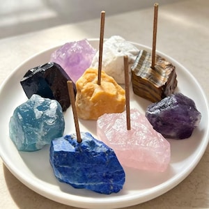 May include: A collection of eight raw, unpolished mineral stones arranged on a white plate. Four of the stones feature thin, vertical incense sticks inserted into their centers. The stones display various colors including blue, purple, yellow, white, and brown.