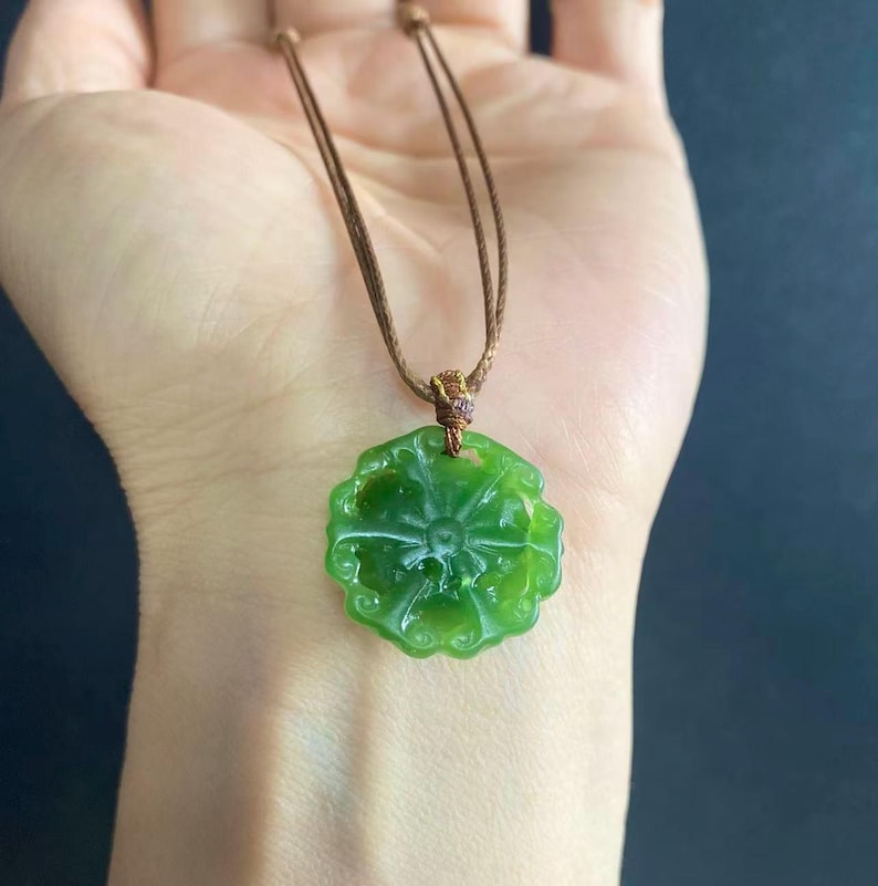 May include: A green jade pendant necklace with a floral design. The pendant is round with carved details and is suspended from a brown cord. The necklace is displayed against a neutral background.
