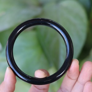 May include: A smooth, circular bangle bracelet with a polished, solid black finish. The bangle features a simple, continuous band design with a rounded profile, showcasing a deep, uniform black color throughout the entire circumference of the piece.