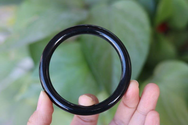 May include: A smooth, circular bangle bracelet with a polished, solid black finish. The bangle features a simple, continuous band design with a rounded profile, showcasing a deep, uniform black color throughout the entire circumference of the piece.