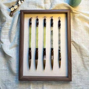 Op de afbeelding: Een ingelijste set van vijf kalligrafie penselen. Elke penseel heeft een donkerbruin handvat, een lichtgroene schacht en een puntige penseelpunt. De penselen worden in een houten frame tegen een witte achtergrond getoond.