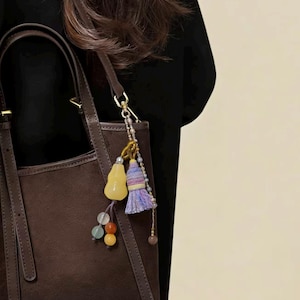 Könnte beinhalten: Eine braune Wildleder-Tragetasche mit passendem Griff und Riemen. Die Tasche ist mit einem dekorativen Anhänger mit einer gelben Kalebasse, bunten Perlen und einer Quaste verziert. Die Tasche hat eine neutrale Farbe und ein schlichtes Design.