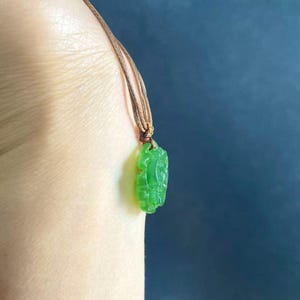 May include: A vibrant green jade pendant carved with a detailed design, suspended from a brown cord necklace. The pendant is a bright, translucent green, and the cord is knotted at the top.