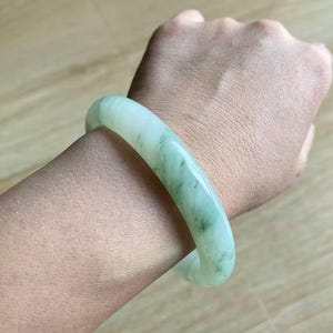 Könnte beinhalten: Ein Jade-Armreif mit glatter, polierter Oberfläche. Das Armband hat eine hellgrüne Farbe mit dunkleren grünen Adern. Der Jade ist durchscheinend und das Armband ist rund.