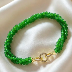 May include: A multi-strand bracelet composed of small, round, vibrant green beads. The bracelet features a circular gold-toned clasp encrusted with small, clear, sparkling stones that connect the two ends of the beaded strands together.